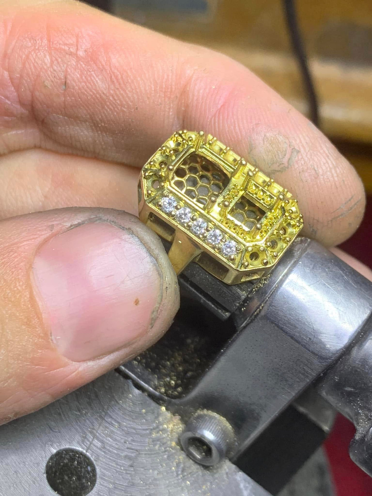 Close-up of an unfinished geometric jewellery setting in a bench clamp, with open stone seats and a partially set row of diamonds.