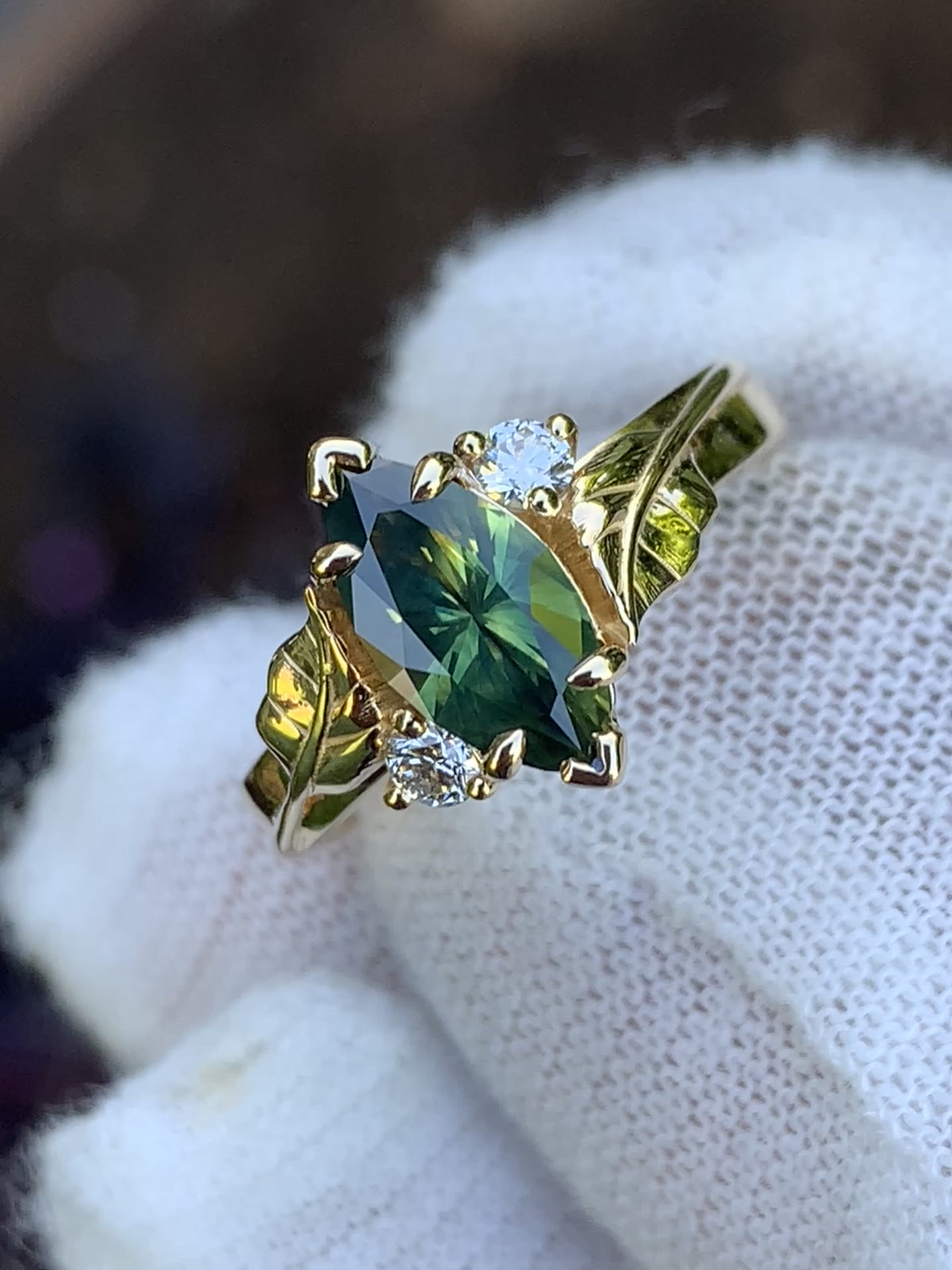 Close-up of a green and yellow sapphire ring with gold leaf detailing and two round white accent stones, held in a white glove.