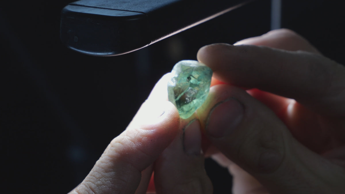 A pale blue-green rough sapphire held between fingertips beneath a bright inspection light, revealing its internal features.