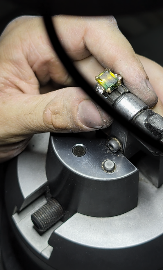 Jeweller’s hands steadying a green and yellow sapphire ring in a bench clamp during setting work.