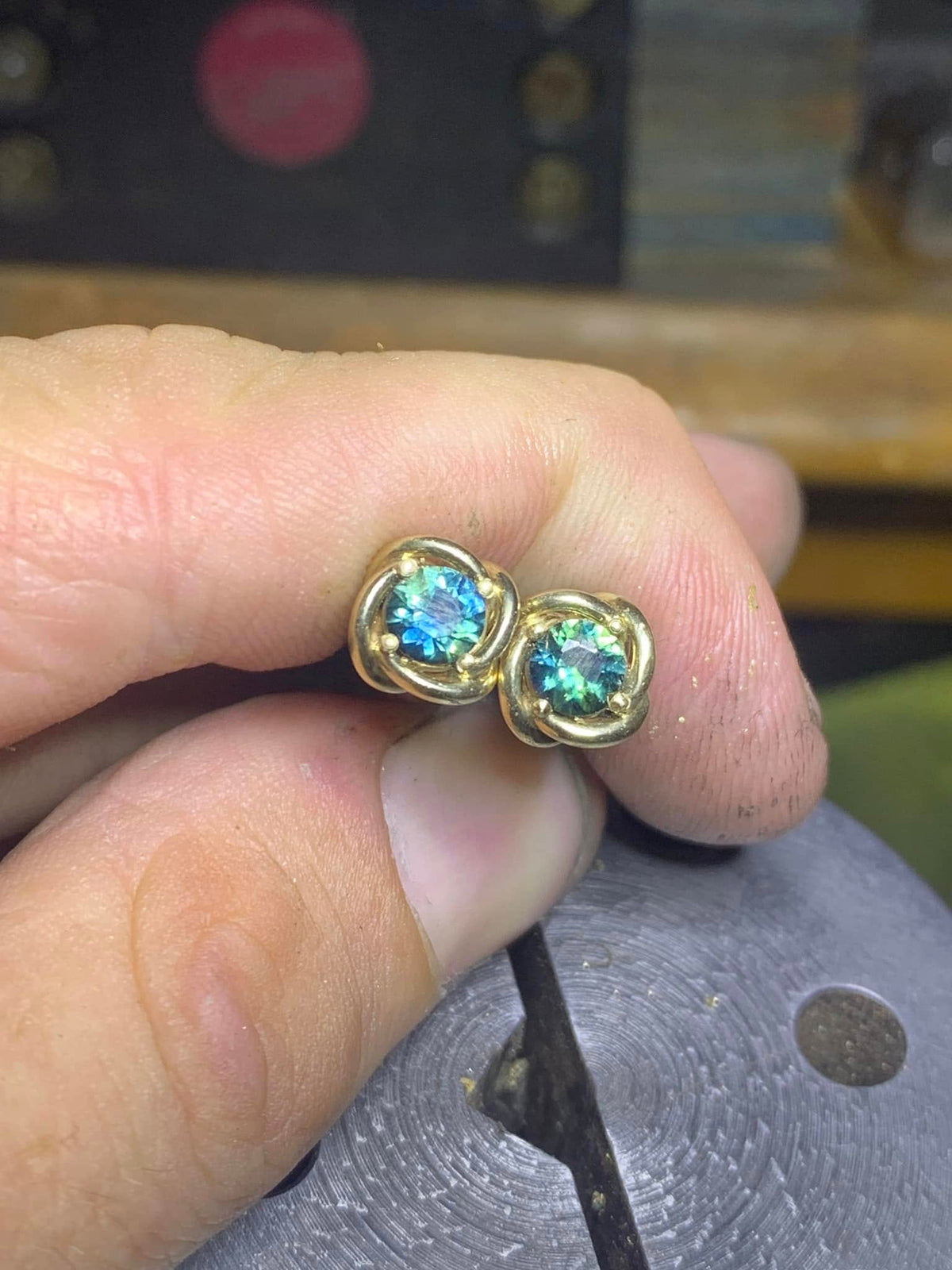 A pair of blue-green sapphire stud earrings with curved metal surrounds, held above a jeweller’s bench clamp.