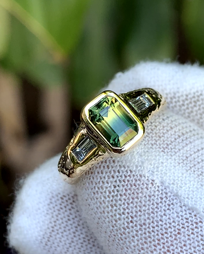 Rectangular green and yellow sapphire in a bezel setting, flanked by rectangular white accent stones and decorative shoulders.